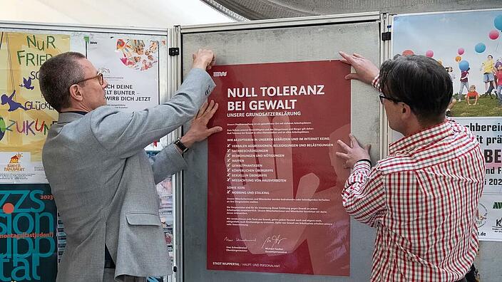 Zwei Personen hängen ein Plakat mit dem Titel "Null Toleranz bei Gewalt" in der Stadtverwaltung Wuppertal auf.