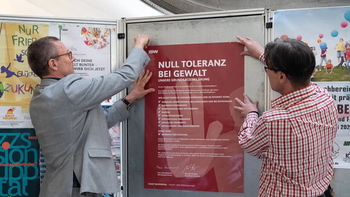 Zwei Personen hängen ein Plakat mit dem Titel "Null Toleranz bei Gewalt" in der Stadtverwaltung Wuppertal auf.