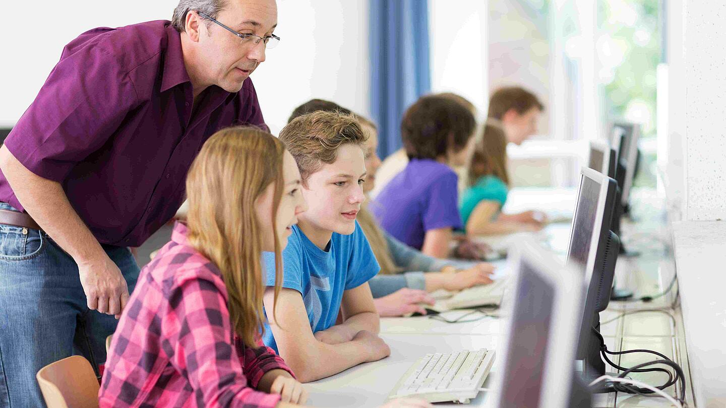 Digitale Schule: Schüler und Schülerinnen arbeiten an Computern, im Hintergrund der Lehrer.